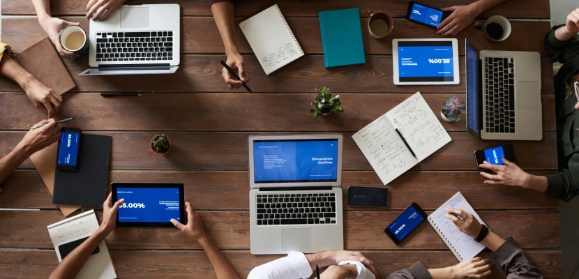 Mitarbeitende sitzen zu einem Projektmanagement Meeting mit Tablets, Laptops und Smartphones an einem Tisch.
