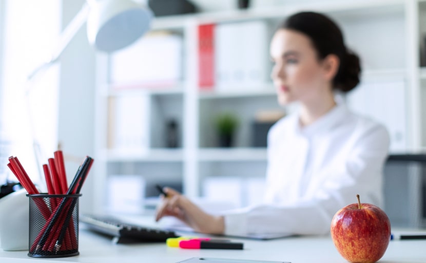 Eine Mitarbeiterin sitzt an einem gut beleuchteten Arbeitsplatz, mit einem Apfel auf dem Schreibtisch.